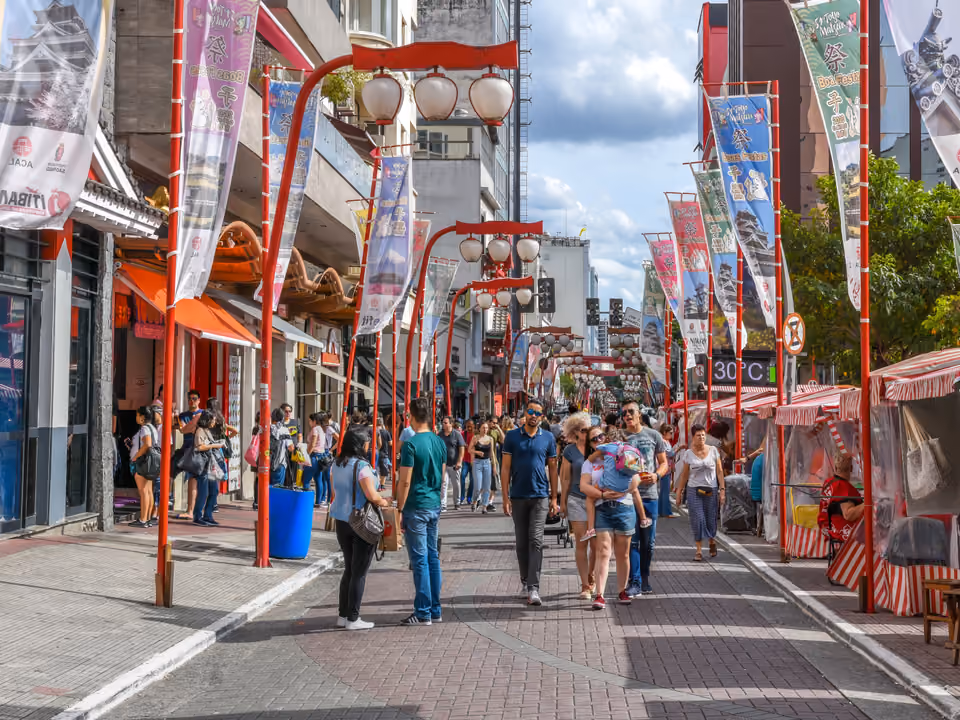 Rua do bairro da Liberdade, em São Paulo, com os postes-lanterna vermelhos (suzuran-tō), as bandeirolas de festival japonês e as barracas listradas da feira, com movimento de pedestres