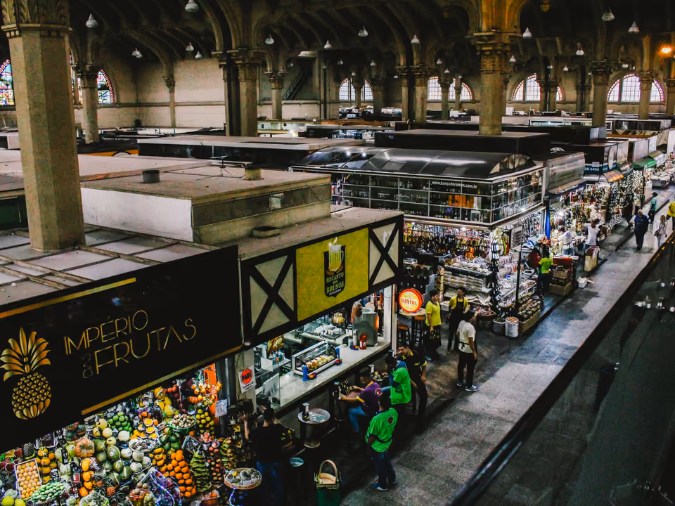 Interior do Mercado Municipal de São Paulo visto do mezanino, as arcadas com colunas e os vitrais em arco sobre o labirinto de boxes, com bancas de fruta em primeiro plano