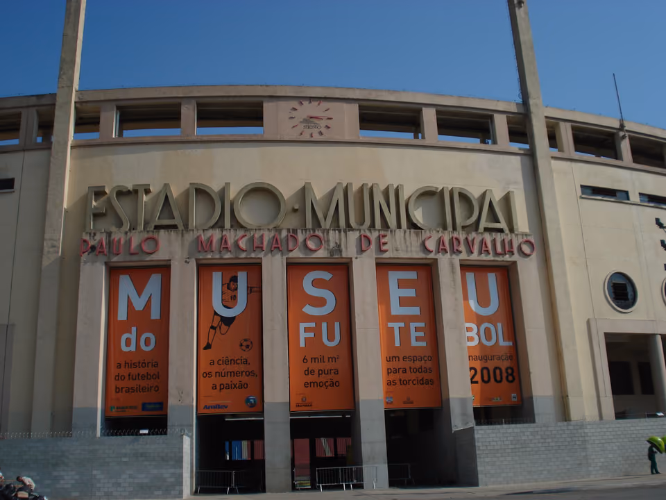 Fachada art déco do Estádio do Pacaembu em São Paulo, com o nome gravado na pedra e os grandes cartazes laranja do Museu do Futebol sobre as entradas em arco