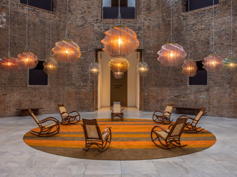 Salão de tijolo aparente da Pinacoteca de São Paulo, com as janelas em vão escuro nas paredes altas e uma instalação de luminárias suspensas e cadeiras de balanço no piso